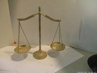 Front view of an ornamental metal balance scale with two hanging pans, displayed on a white surface against a plain background.