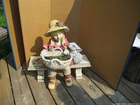 Full front view of outdoor girl figurine sitting on a stone-look bench, holding a basket with a lamb figure beside her.