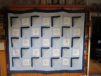 Full view of the quilt hanging, showing its squared patchwork pattern with white and floral blocks framed in dark fabric segments and bordered by light blue checkered fabric.