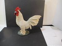 Front and side view of a large white resin rooster figurine with red comb and wattles, standing on a green base.