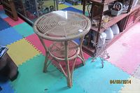 Round rattan table with glass top on colorful foam mat flooring, showing woven rattan patterns and angled leg supports.