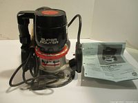 Sears Super Router front view showing black and red router with power cord and clear base plate, placed beside its user manual.