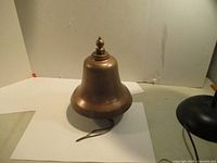 Front and side view of solid brass bell on display, showing overall shape and decorative top finial.