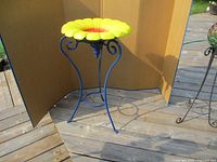 Full view of sunflower bird bath showing the yellow and orange flower basin and blue metal stand on wooden deck with cardboard backdrop.