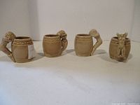Front view of four beige ceramic shot glasses set in a row showing sculpted figure handles.
