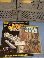 Photo of two vintage comics stacked, showing the Battlestar Galactica Marvel Super Special cover with the official ship art and Star Wars Marvel Super Comics Group cover featuring Star Wars characters in an action scene.