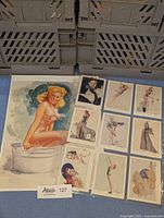 Photo showing a large magazine cutout of a blonde pin-up woman sitting on a barrel accompanied by eight smaller pin-up cards in a grid.