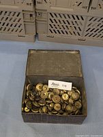 Metal box filled with numerous brass buttons, showing the variety of sizes and designs from a top perspective.