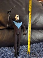 Nightwing figure standing with bow accessory held up and measuring tape showing height about 12 inches.