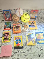 Overview photo of assorted vintage collectibles on countertop featuring trading card wrappers, signed baseballs, stickers, and toy figure.