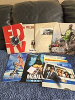 Seven assorted vintage movie press releases displayed on a blue carpet and brown couch, showing various titles and cover designs including Ed TV, Another You, Black Knight, Honeymoon In Vegas, and McHale's Navy.