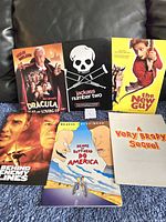 Photo showing six vintage movie press folders arranged on a blue surface against a leather background. Titles visible: Dracula: Dead and Loving It, Jackass Number Two, The New Guy, Behind Enemy Lines, Beavis and Butt-head Do America, A Very Brady Sequel.