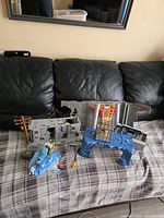 Full view of Batman-themed playsets on a grey and plaid surface on a black leather couch; includes ruined building wall, blue ice platform and pole prop.