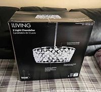 Box of new, unopened Decor Living 3 Light Chandelier showing crystal accent shade and polished chrome finish, held on a couch.