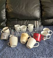 Lot of mixed coffee mugs and clear glass drinkware displayed on blue surface and black sofa backdrop