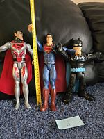 Three DC Comics action figures shown together: two Superman figures and one Batman figure, posed standing upright on a carpeted surface with a measuring tape showing height approx. 12 inches for Superman figures.