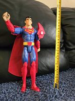 Superman 12-inch action figure standing upright next to measuring tape showing height approximately 12 inches.
