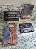 Two Foreigner cassette tapes displayed on a countertop, one upright showing its album case 'Agent Provocateur' and the other case front 'Unusual Heat' with tapes in front of cases.