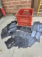 Multiple black plastic garden edging panels laid out next to a red plastic milk crate on a concrete floor in front of a brick wall.