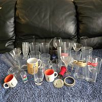 Wide view of various glass drinkware and coffee mugs arranged on carpet in front of black leather couch, showing branded and novelty items