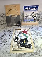 Front view of the lot showing the Beehive redemption card, Eddie Shack card in wooden holder, and Roberto Luongo card on a granite surface.