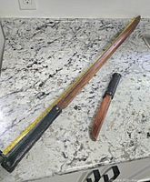 Wooden katana practice sword and wooden dagger placed on marble countertop along with a measuring tape showing size of katana sword.