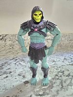 Front view of Skeletor figure showing detailed sculpting, yellow skull face with red eyes, green muscular body, and black and purple armor pieces.