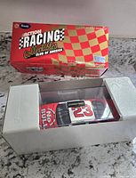 Photo of the packaged 1:24 scale NASCAR die cast car in clear display case with foam inserts and original box showing branding and checkered design.