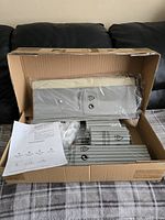 Box opened showing unassembled components of a 6-layer shoe rack including metal rods, plastic parts, and base panels wrapped in plastic along with instruction sheet.