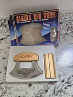 Top view showing the ulu knife blade with eagle engraving, wooden handle, and wooden stand inside white insert tray, with original box packaging behind.