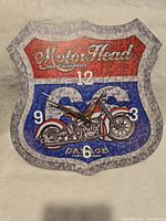 Front view shot of Motor Head Parts & Service wall clock shaped like a shield with motorcycle illustration and numbers.