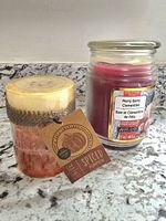 Two candles on a granite countertop. One is layered cream and orange with a burlap band and label reading 'Spiced Pumpkin'. The other is a glass jar candle labeled 'Merry Berry Clementine' with dark pink wax inside.