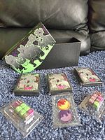 Photo showing black and green Pokemon trading card storage box with lid off, three sealed packages of penny sleeves with Pokemon artwork, and multiple sets of colorful dice and plastic tokens.