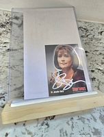 Autograph card signed by Lorraine Bracco as Dr. Jennifer Melfi, displayed in front of a white sheet on a countertop with a wooden holder.