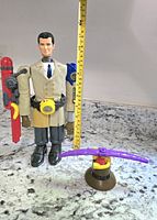 Photo of Inspector Gadget action figure with yellow measuring tape showing height approx 10 inches. Wearing beige trench coat, blue tie, black shoes, with red jetpack and purple helicopter gadget accessory.