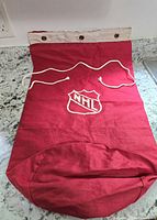 Full view of vintage red canvas NHL equipment bag with white NHL shield logo and thick white rope drawstring, laid flat on countertop.