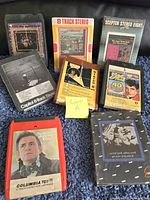 Photo showing eight vintage 8-track stereo music tapes in original cases laid out on a dark cloth surface.