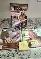Three vintage WWF/WWE magazines on a granite countertop. One magazine shows Steve Austin and Big Bossman on cover, one is WWF Magazine 1997, and the third is The Wrestler with Kurt Angle on cover.