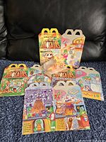 Lot arranged on blue carpet showing seven colorful McDonald's Dinosaurs Happy Meal boxes with cartoon characters and puzzles, all unopened and in new condition.