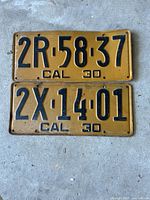 Two 1930 California vintage license plates stacked one above the other on a concrete surface showing embossed black text '2R 58 37 CAL 30' and '2X 14 01 CAL 30' on yellow background with wear and rust on edges.
