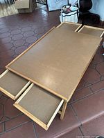 Wooden puzzle table with foldable legs and three pull-out drawers in light brown finish placed on floor.