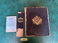 Two books on a green surface, the larger with a damaged brown cover with a gold Masonic symbol and a smaller booklet titled The Master Mason.