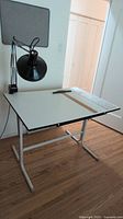 Photo of drafting table with adjustable metal legs, white top, attached black adjustable lamp, wooden and plastic rulers, and cutting mat on wooden floor.