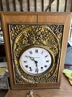Front view of the grandfather clock showing the wooden case and ornate brass decorative face surrounding the round white clock face with Roman numerals and black decorative hands.