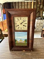 Front view of rectangular wooden wall clock showing clock face with Roman numerals and painted glass panel below depicting a building with trees