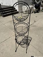 Full front view of black metal 3-tier fruit stand positioned outdoors on concrete showing stand height and structure.