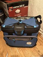 Blue Titan cooler with zipperless ColdLok closure, front zippered pocket and shoulder strap, shown with stoneware bake set box in background.