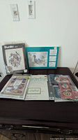 Overview of multiple embroidery and cross stitch kits laid out on table including Bucilla and Design Works kits, some sealed and some opened.