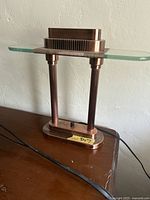 Desk lamp sitting on a wooden surface showing clear visibility of lamp base, glass shelf, and black ventilated lamp housing.