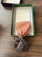 Silver-plated medal in green-lined box, orange ribbon attached, front view shows male athlete in relief